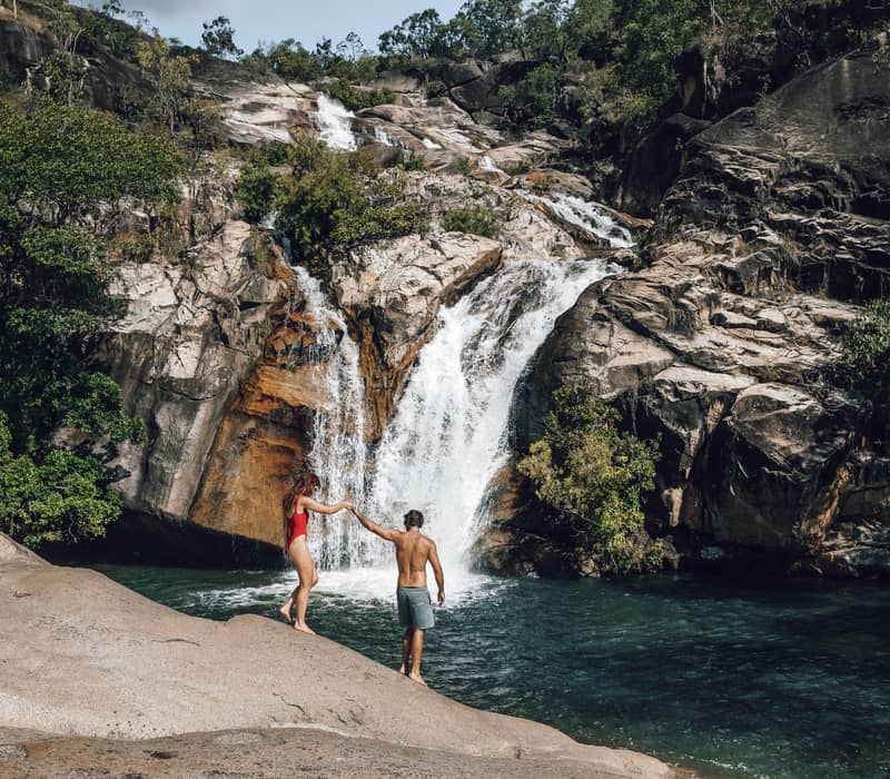 A man and woman in swimwear relaxing on large granite rocks by a scenic waterfall at Emerald Creek Falls, Mareeba.