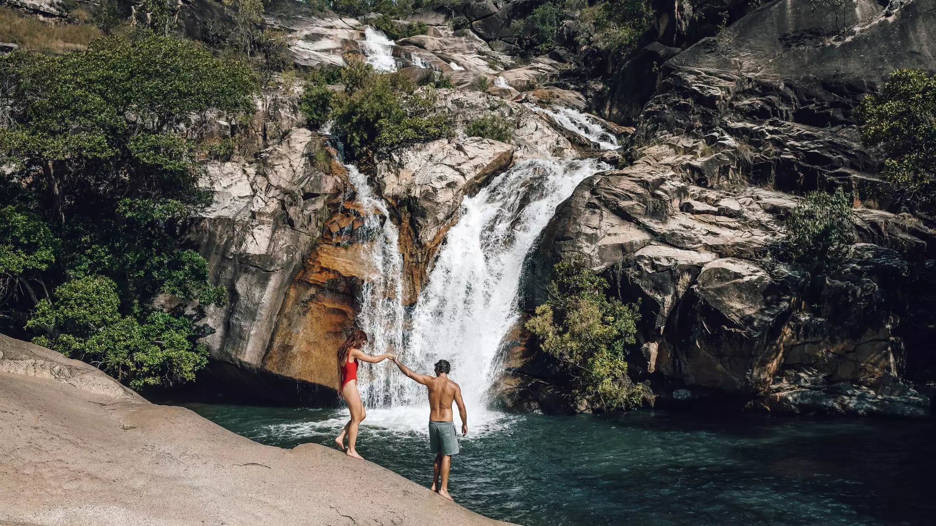 A man and woman in swimwear relaxing on large granite rocks by a scenic waterfall at Emerald Creek Falls, Mareeba.