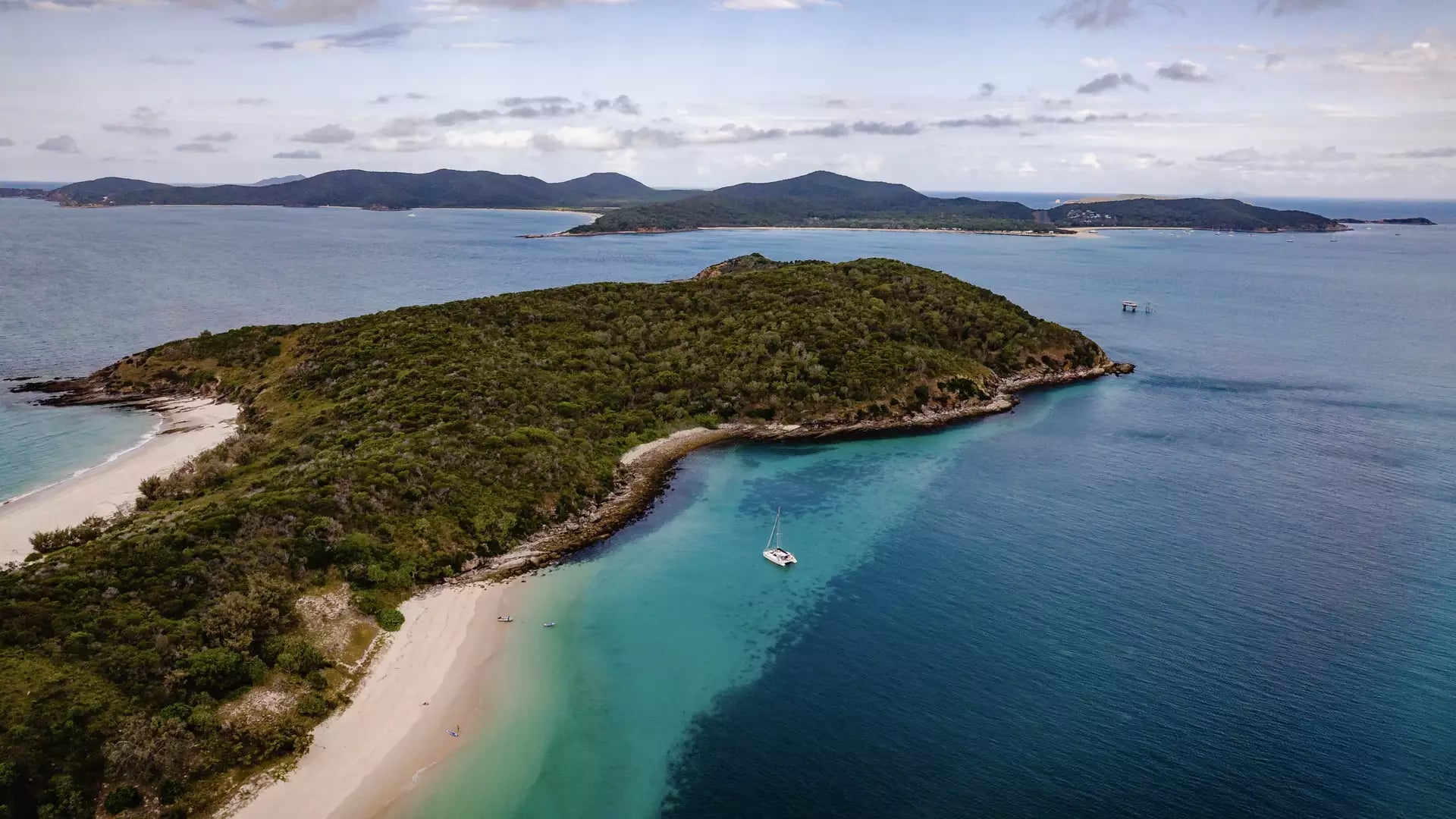 Aerial view of a white sailing boat in a clear blue bay next to a forested island and sandy beach.