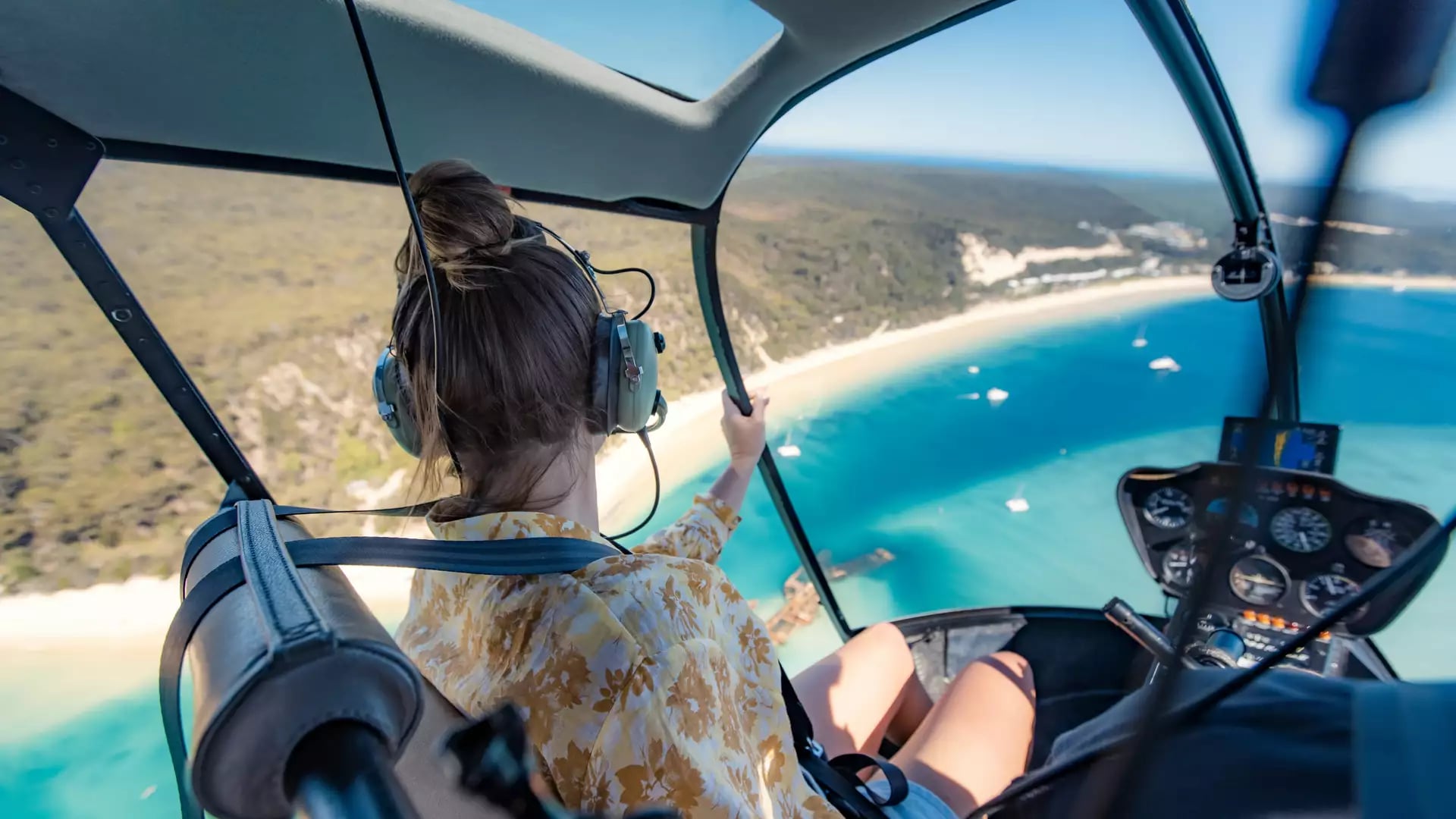 A view from inside the cockpit of a helicopter flying over the turquoise waters and rusted Tangalooma Wrecks alongside the massive white sand dunes of Moreton Island in Queensland.
