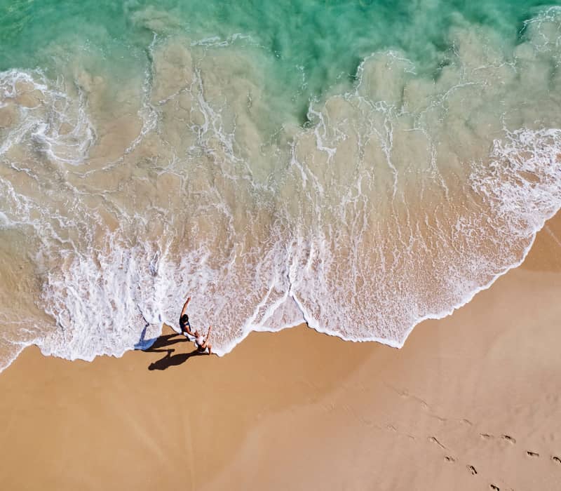 A vertical top-down aerial photograph showing the shallow, white-foaming surf of a beach. The water is a pale turquoise, meeting smooth golden-tan sand.