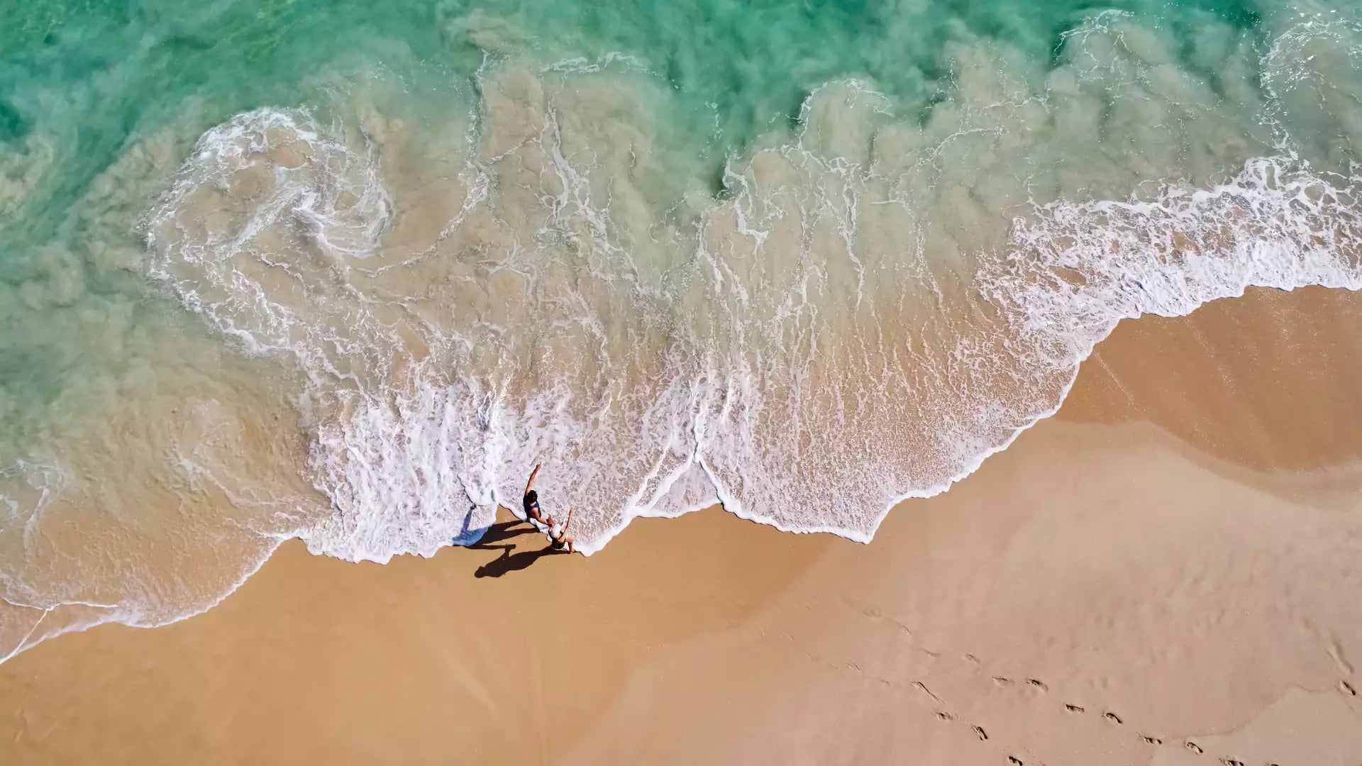 A vertical top-down aerial photograph showing the shallow, white-foaming surf of a beach. The water is a pale turquoise, meeting smooth golden-tan sand.