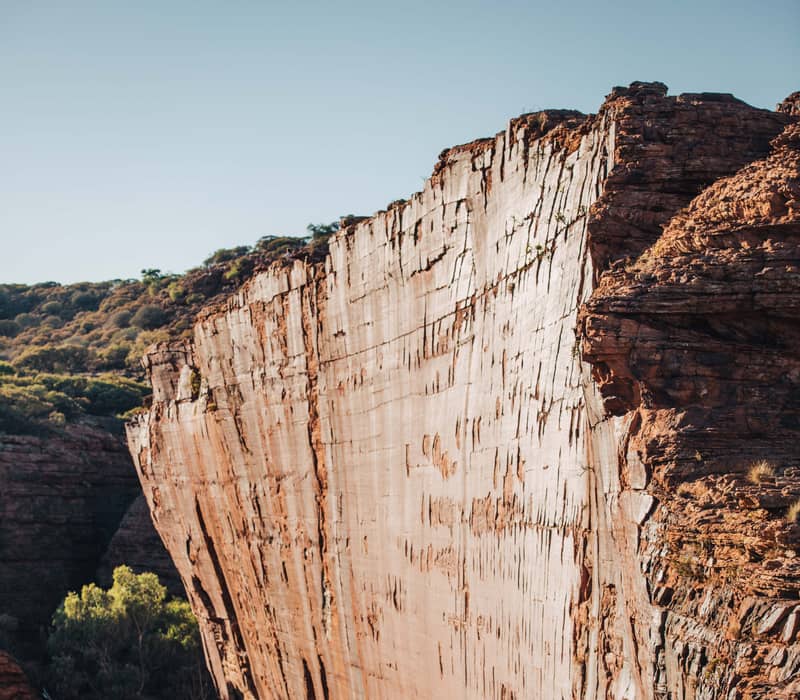 The edge of a massive, vertical red rock cliff at Kings Canyon, with weathered sandstone domes in the background under a soft blue sky.