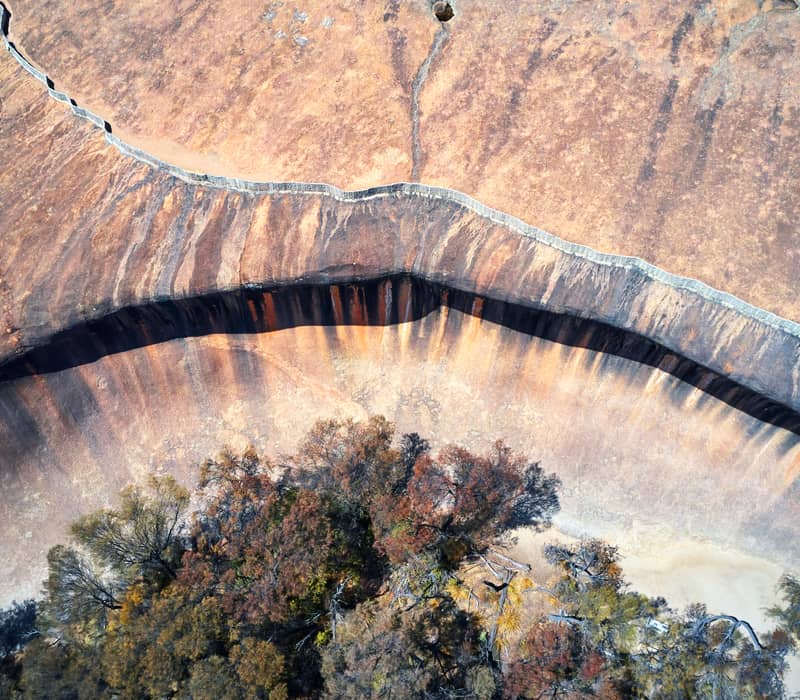 An aerial top-down view of the iconic curved formation of Wave Rock surrounded by the dense green scrub of the Wheatbelt in Hyden.