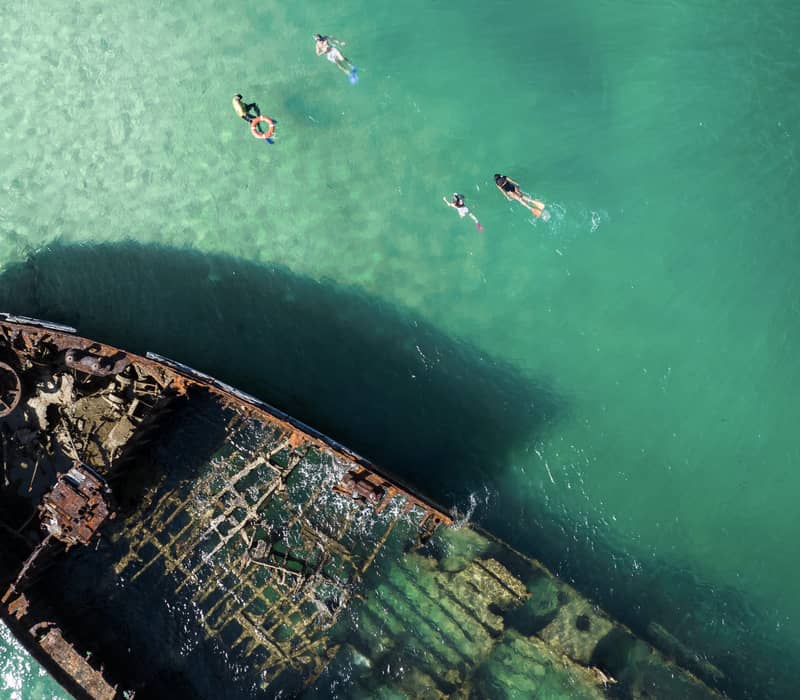 From a top-down aerial view, snorkellers swim through the clear blue water surrounding the rusted hulls of the Tangalooma Wrecks on Moreton Island, Queensland.