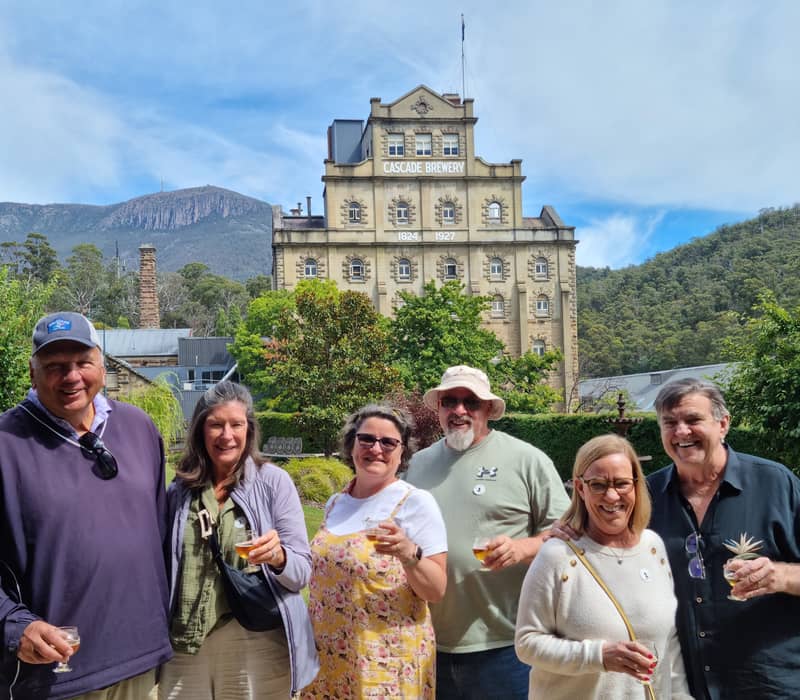 Six people smiling and holding beer tasting glasses in front of the historic stone facade of Cascade Brewery in Tasmania.