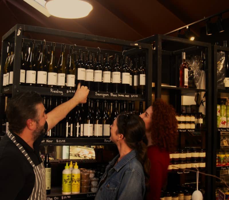 A man in an apron pointing to wine bottles on a dark shelf for two women in a gourmet food and wine shop in Hobart.