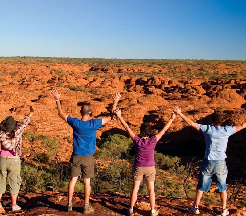 Four travelers with their arms raised in the air standing on a rocky ledge overlooking a vast landscape of orange rock formations.