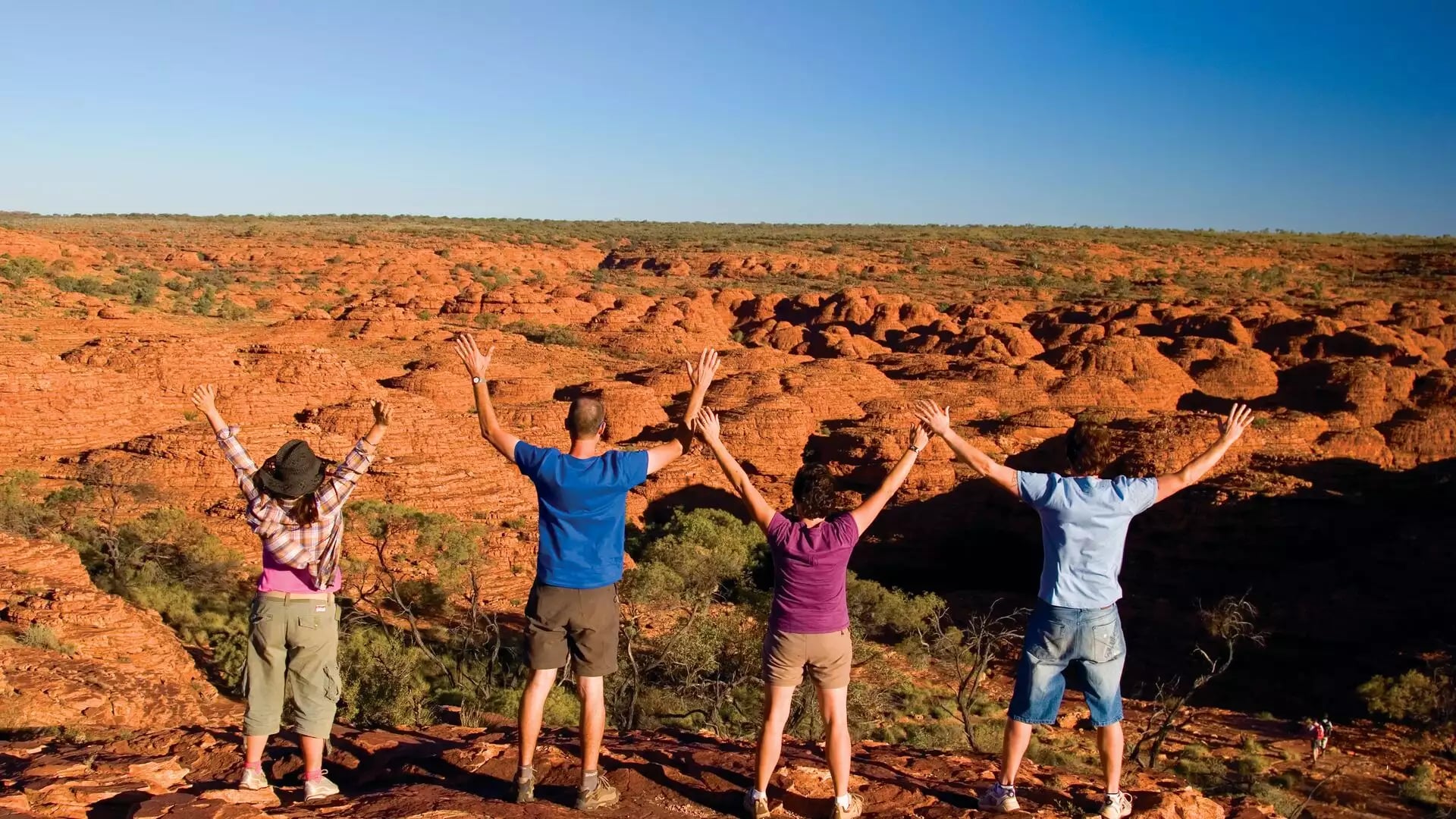 Four travelers with their arms raised in the air standing on a rocky ledge overlooking a vast landscape of orange rock formations.