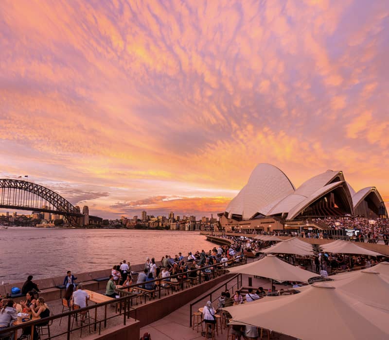 Vibrant sunset over the Sydney Opera House and Harbour Bridge with people enjoying drinks by the water.