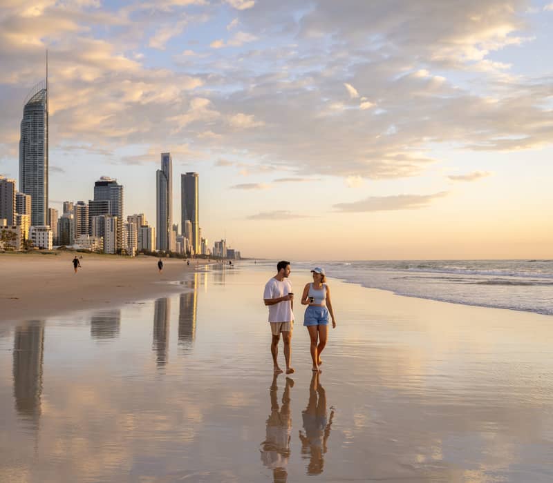A couple walking along the wide, reflective shoreline of Surfers Paradise beach with high-rise buildings under a soft morning sky.