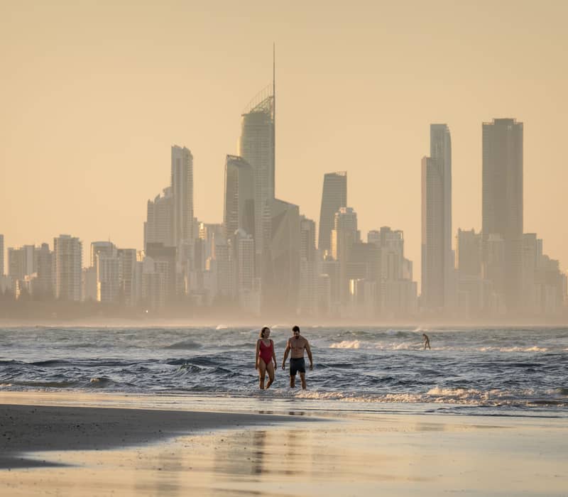 Aerial view of many surfers in the ocean at Currumbin Alley with a large rock formation and city skyline on the horizon.