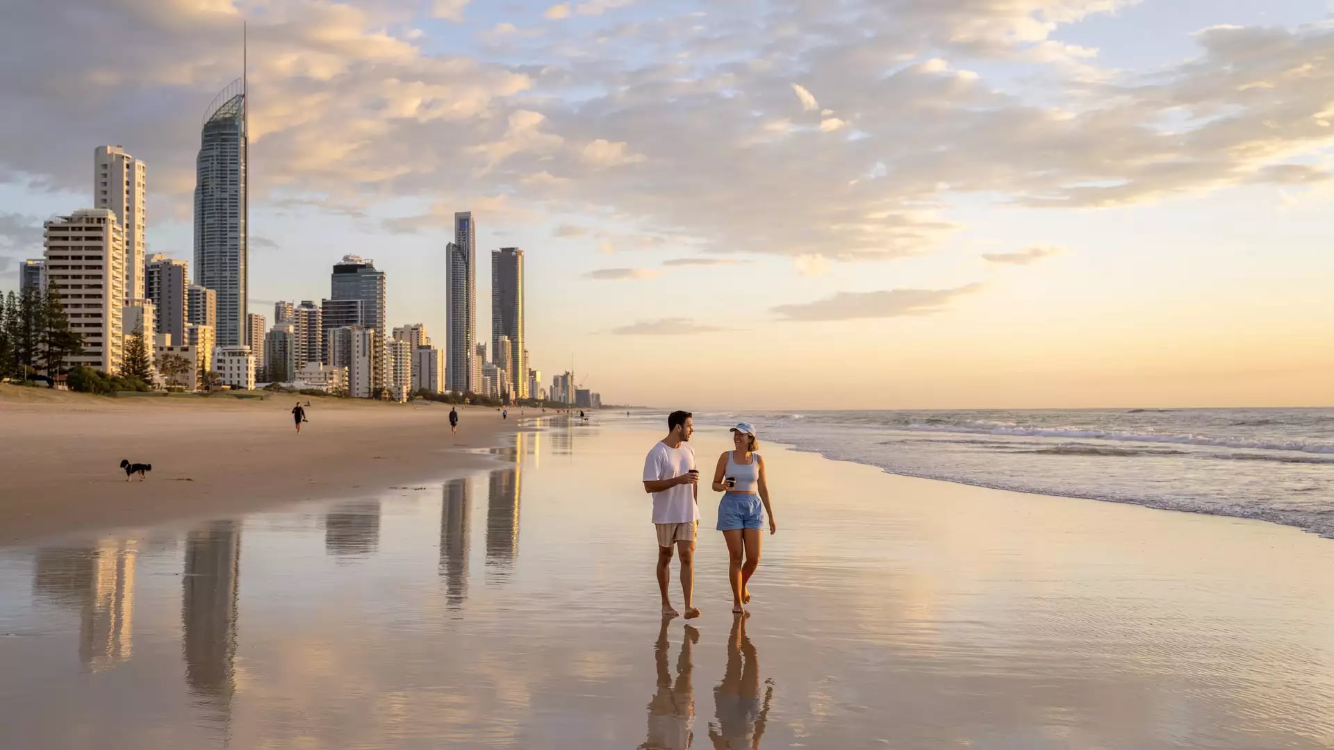 A couple walking along the wide, reflective shoreline of Surfers Paradise beach with high-rise buildings under a soft morning sky.