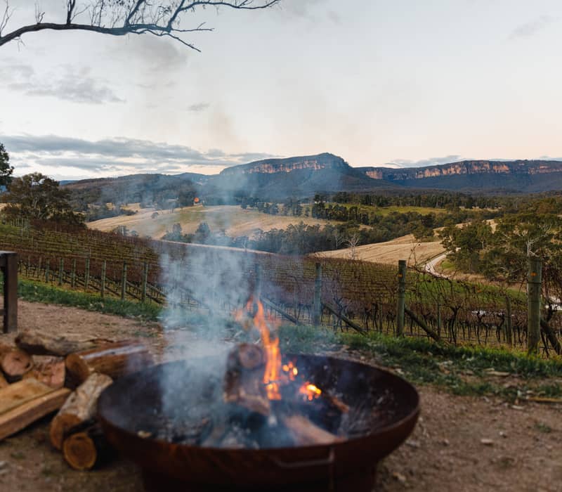 A smoking wood fire pit overlooking rolling green vineyard rows and distant sandstone cliffs in the Blue Mountains, NSW.