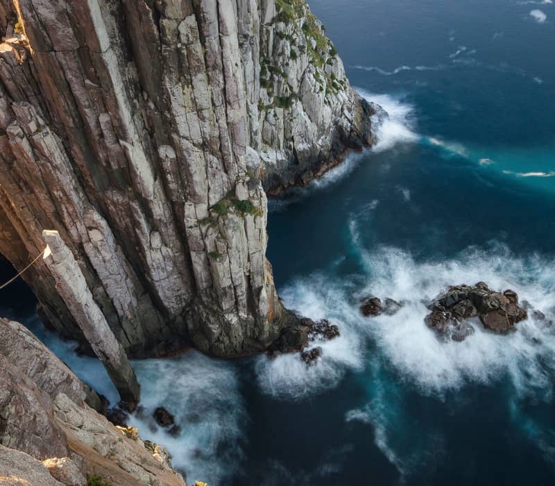 A dramatic top-down view looking directly over the edge of vertical dolerite sea cliffs at Cape Pillar, featuring the iconic "Totem Pole" and "Candlestick" rock pinnacles surrounded by churning white water from the Southern Ocean