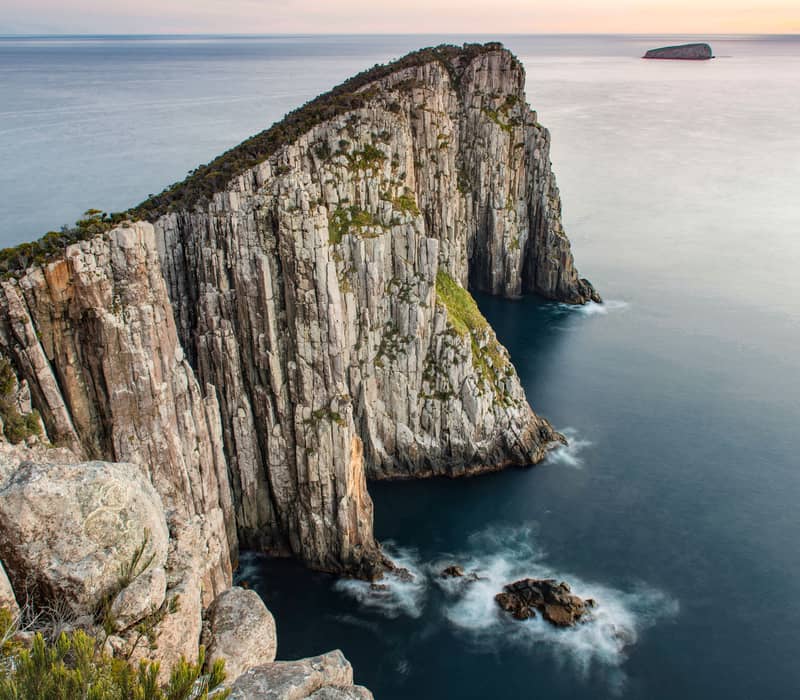 Dramatic landscape view of the jagged dolerite sea cliffs at Cape Hauy along the Three Capes Track, showing the "Lanterns" rock formations extending into the calm grey sea under a soft twilight sky