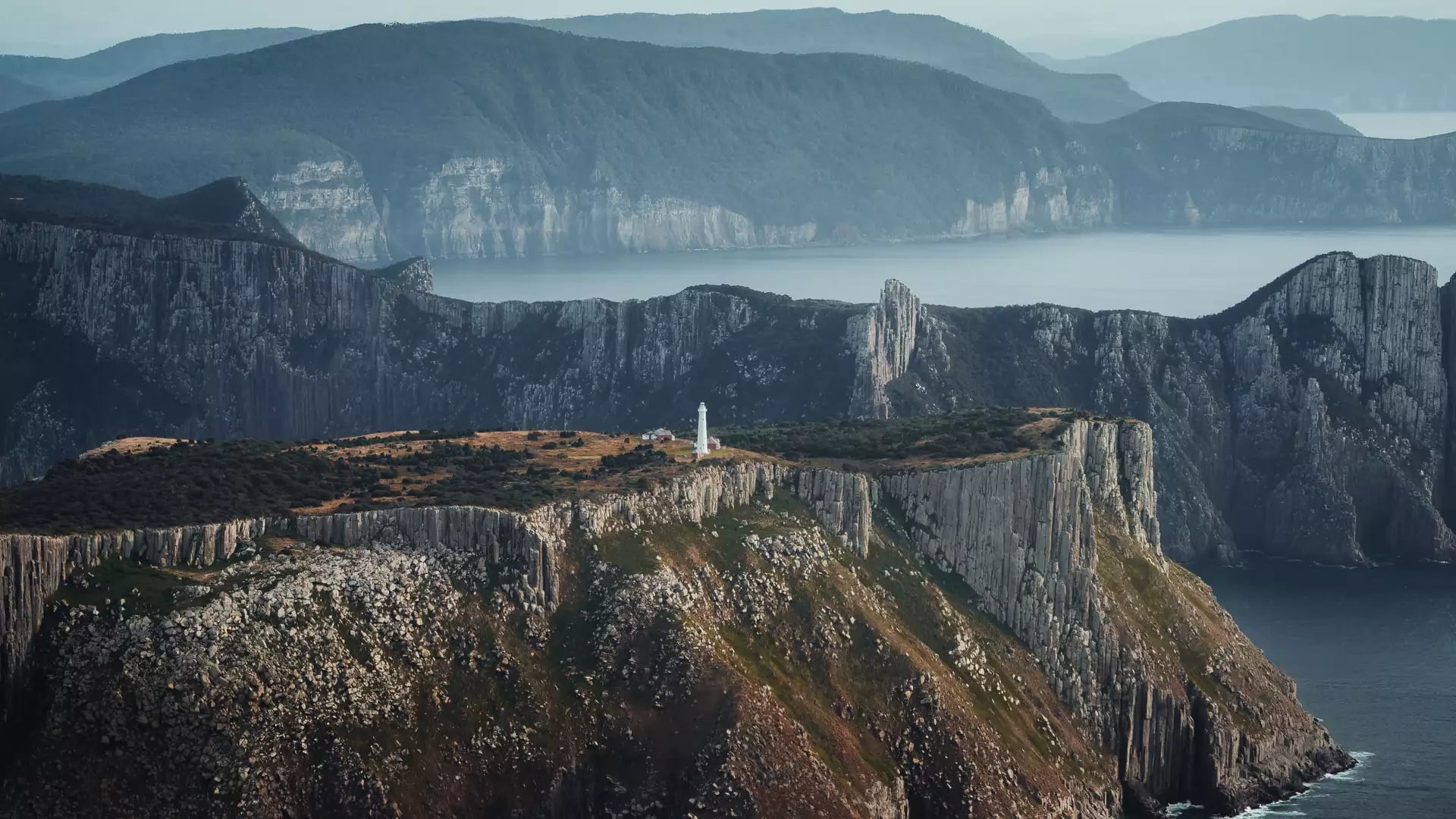 A dramatic landscape view from the summit of "The Blade" on the Three Capes Track, showing the narrow dolerite column formation jutting out into the ocean with Tasman Island and its lighthouse in the background.