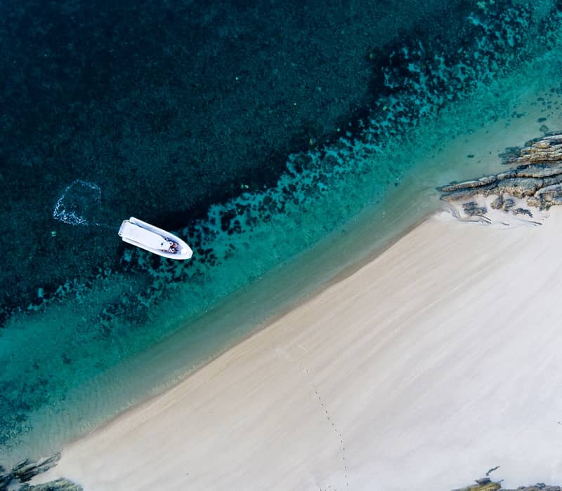 Aerial view of a white boat anchored at a pristine white sand beach with clear turquoise water.