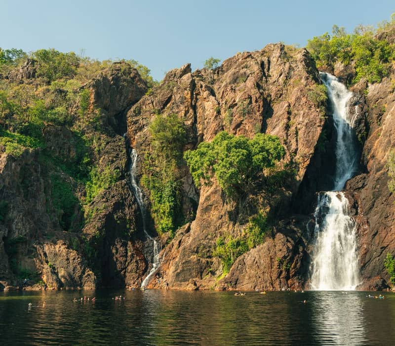 A wide shot of two tall waterfalls flowing down a rocky cliff into a wide dark pool where people are swimming, surrounded by green trees.