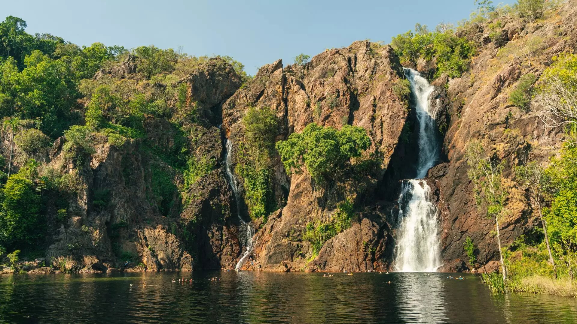 A wide shot of two tall waterfalls flowing down a rocky cliff into a wide dark pool where people are swimming, surrounded by green trees.