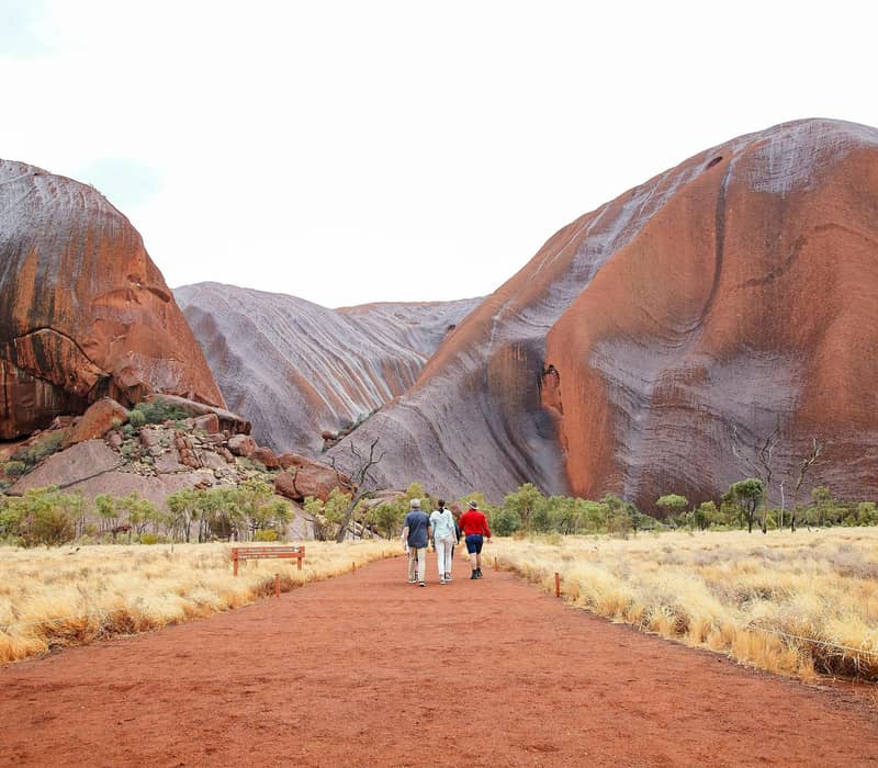 People walking on a red dirt track toward the base of Uluru under a soft sky in Central Australia.