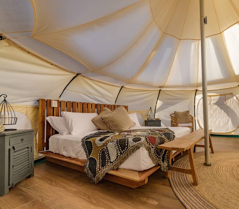 Interior of a glamping tent showing a large bed with a patterned throw, wooden furniture, and woven rugs.