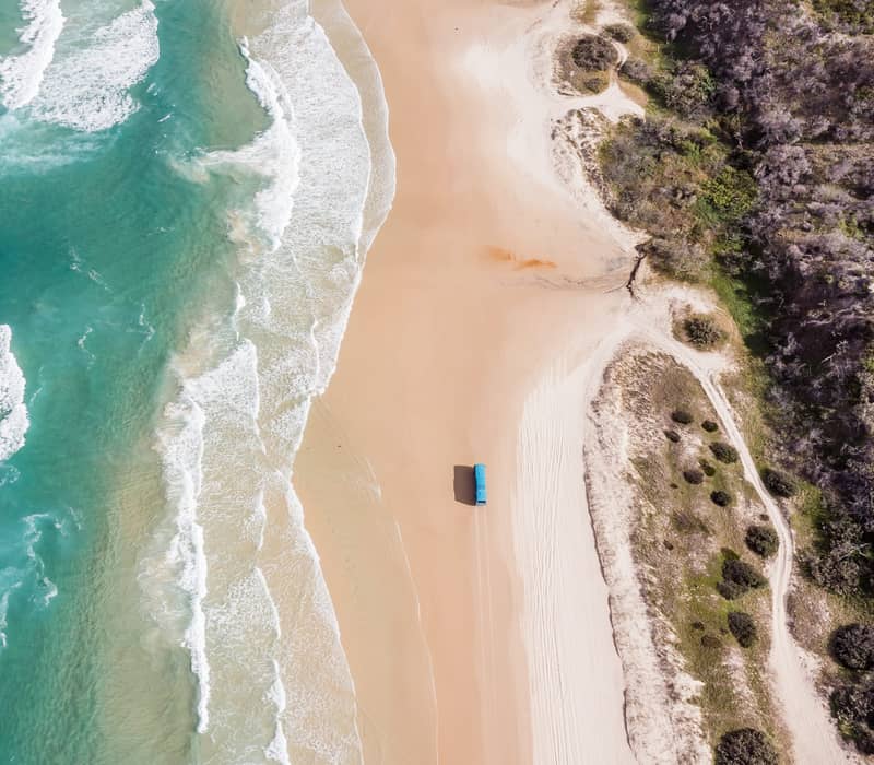 Top-down aerial view of a blue vehicle on a vast beach next to swirling turquoise ocean water.