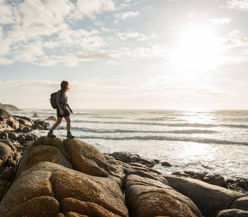 A female hiker with a backpack walks across large orange lichen-covered coastal rocks next to the ocean surf during a bright day in Tasmania.