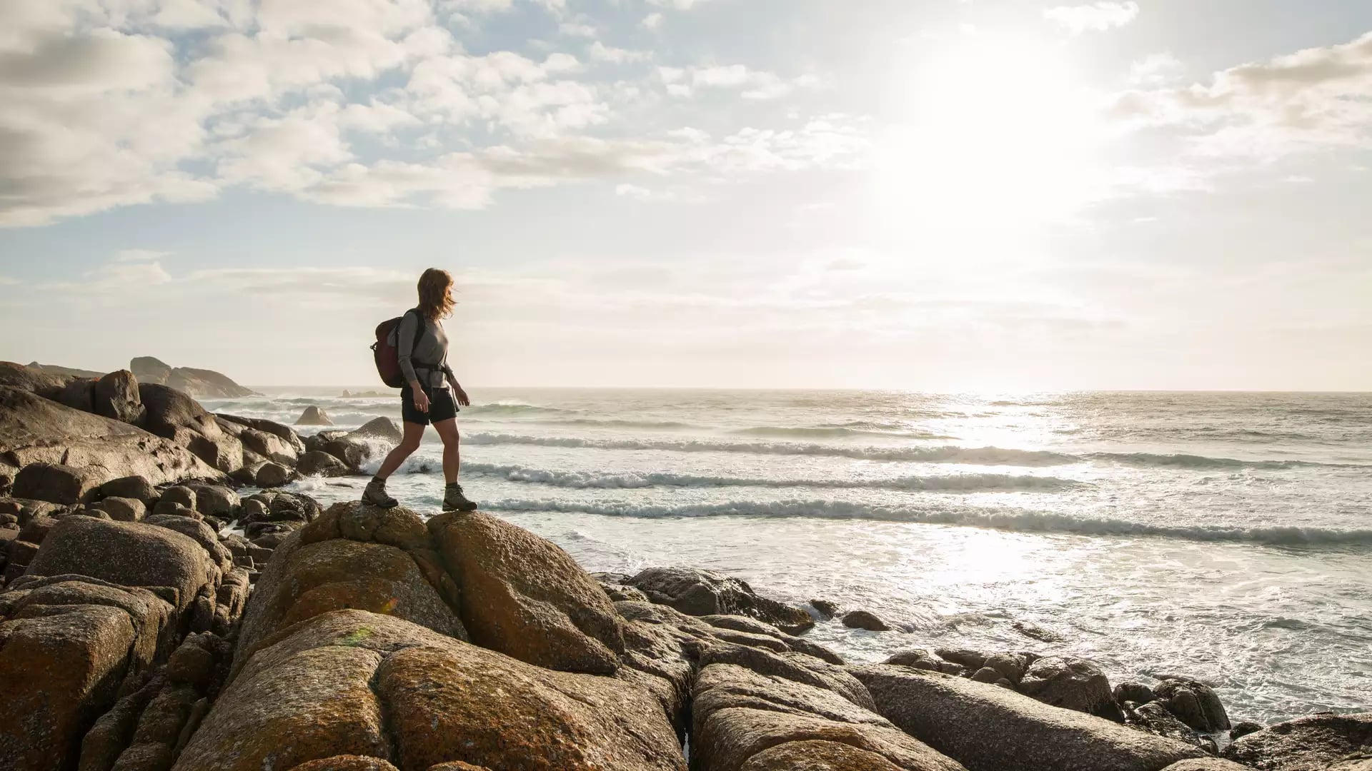A female hiker with a backpack walks across large orange lichen-covered coastal rocks next to the ocean surf during a bright day in Tasmania.