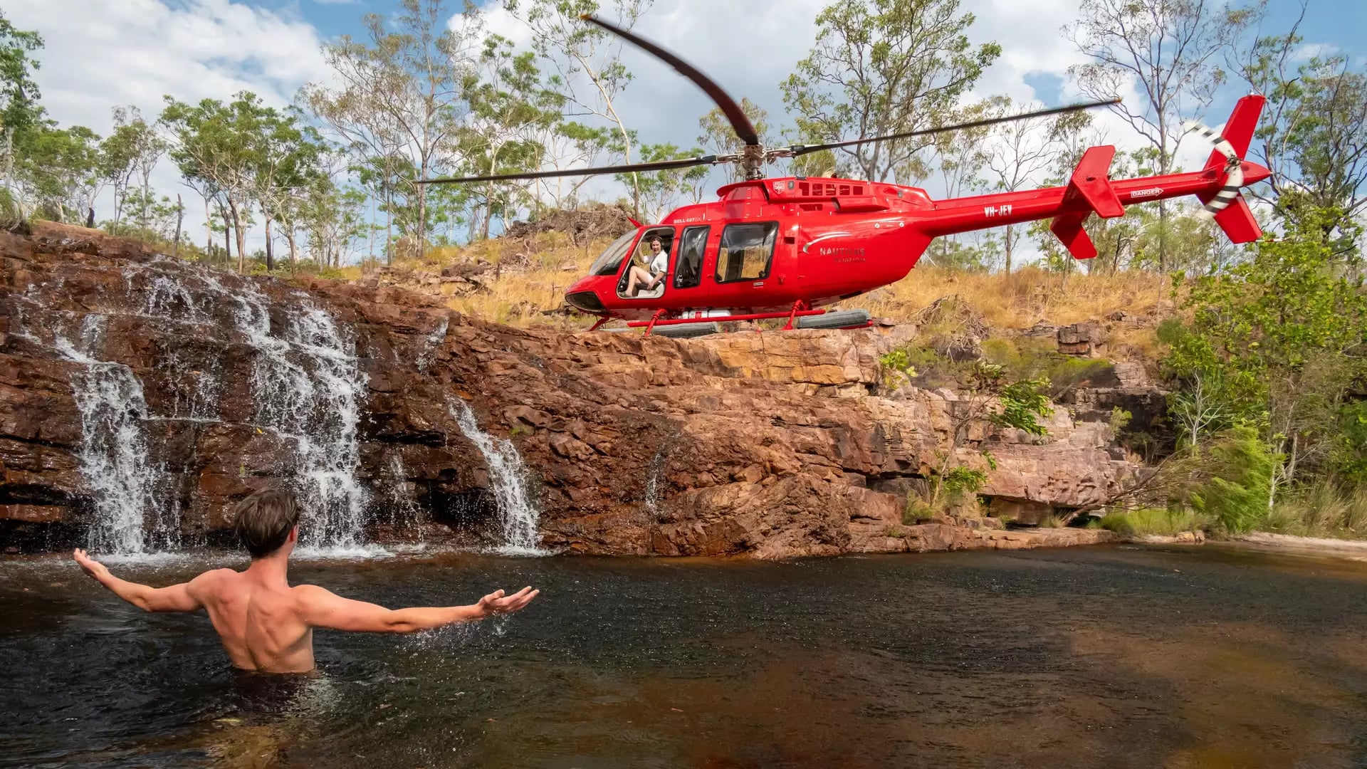 A red helicopter perched on rocks next to a person swimming in a natural waterfall pool in the Northern Territory.