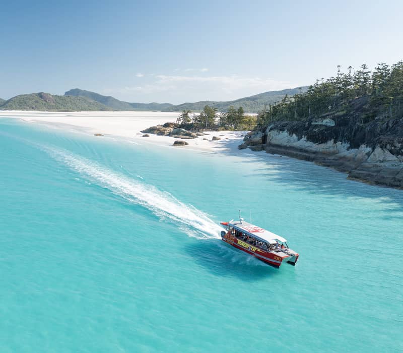Aerial view of a red tour boat cruising past a pristine white sand beach in the Whitsundays.