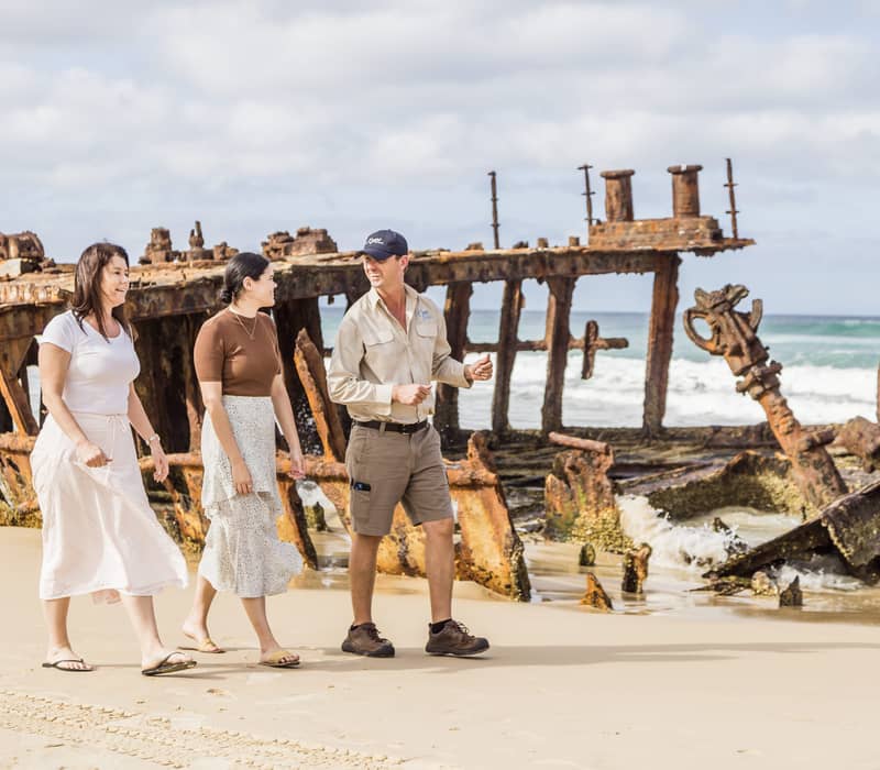 A tour guide and two visitors walking along the sand toward the large, rusted Maheno shipwreck on K'gari.