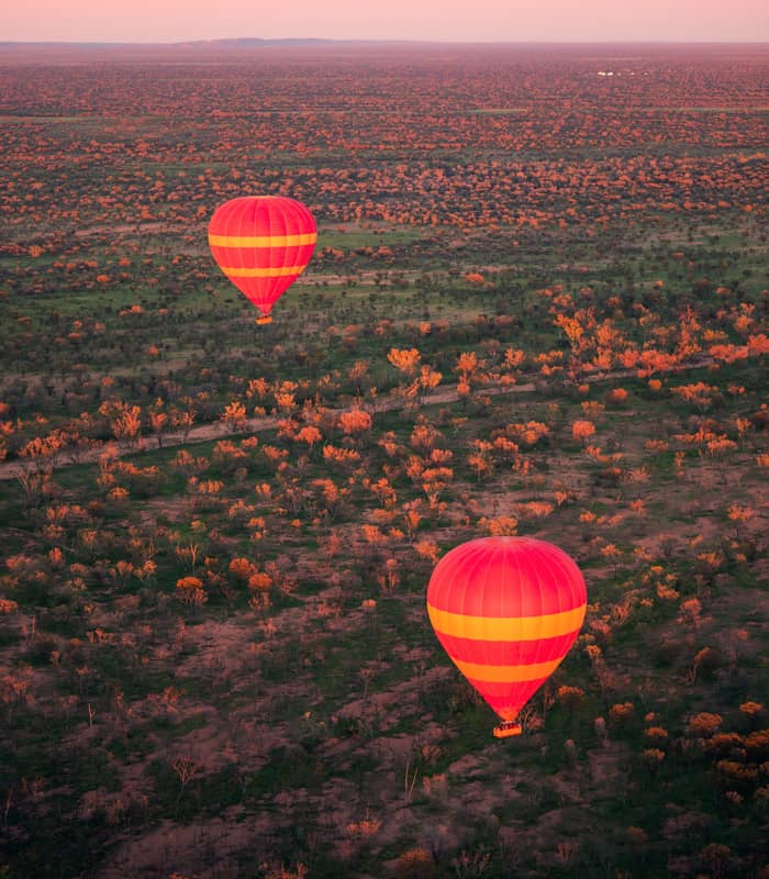 Two vibrant red and orange hot air balloons float gracefully over the vast expanse of the MacDonnell Ranges near Alice Springs at dawn. The early morning light casts a warm, golden glow over the rugged desert landscape and sparse vegetation, offering a serene and bird's-eye perspective of the Northern Territory's iconic Red Centre.