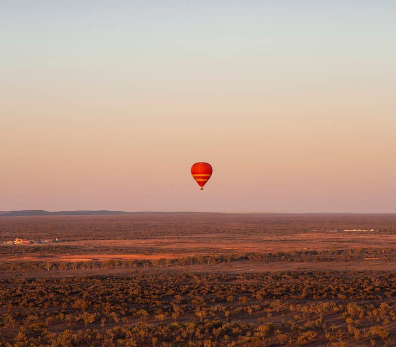 A wide landscape shot at dawn showing a vibrant red and yellow hot air balloon floating above the vast, flat desert plains of the Northern Territory with the rugged blue silhouette of the MacDonnell Ranges on the horizon.