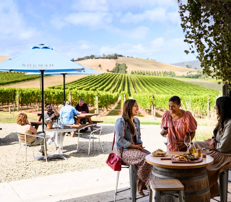 Three women sitting at a table outdoors at a winery, laughing and drinking wine with views of a green vineyard.
