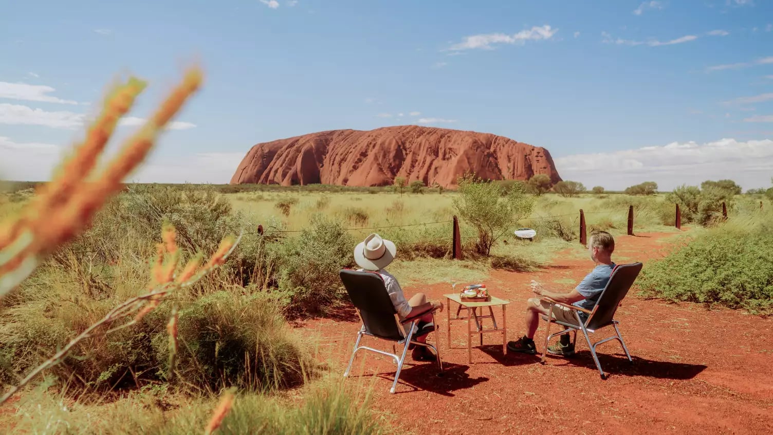 Two people sitting in camping chairs with a small table of food overlooking Uluru in the desert.
