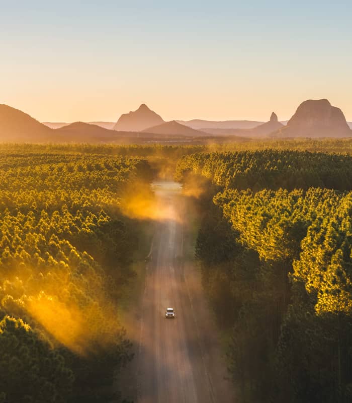 A high-angle aerial view of a white car driving toward the base of Mount Ngungun, highlighting the intersection of forestry and ancient volcanic peaks.