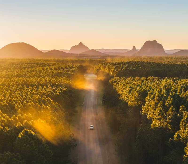 A high-angle aerial view of a white car driving toward the base of Mount Ngungun, highlighting the intersection of forestry and ancient volcanic peaks.