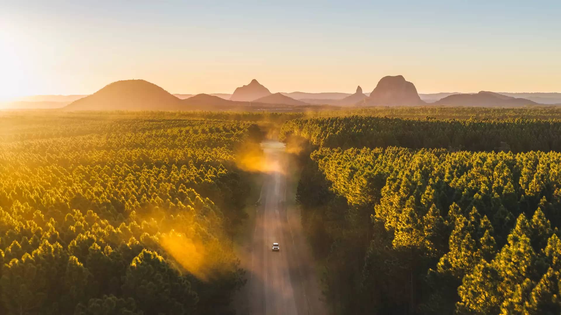 A high-angle aerial view of a white car driving toward the base of Mount Ngungun, highlighting the intersection of forestry and ancient volcanic peaks.