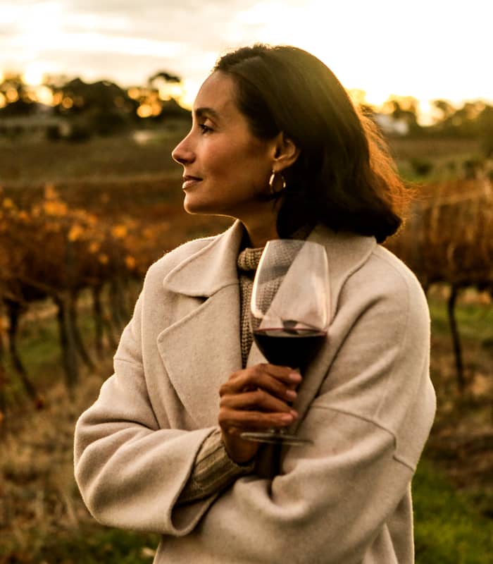 A woman in a cream coat holding a glass of red wine in a Barossa Valley vineyard at sunset.