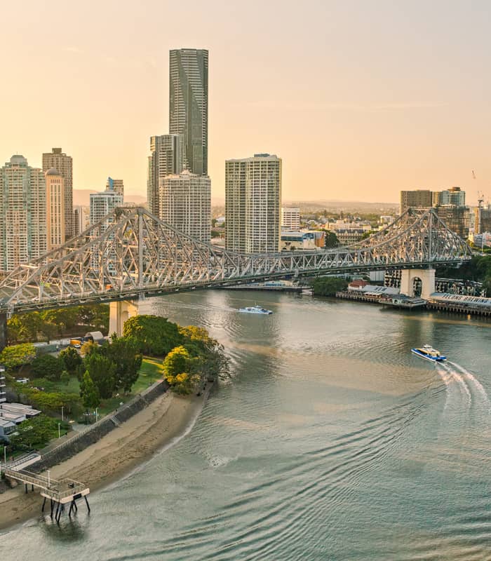 An aerial view of a wide river with several boats leaving white wakes, passing under a large steel bridge toward a city of tall buildings under a hazy yellow sky.