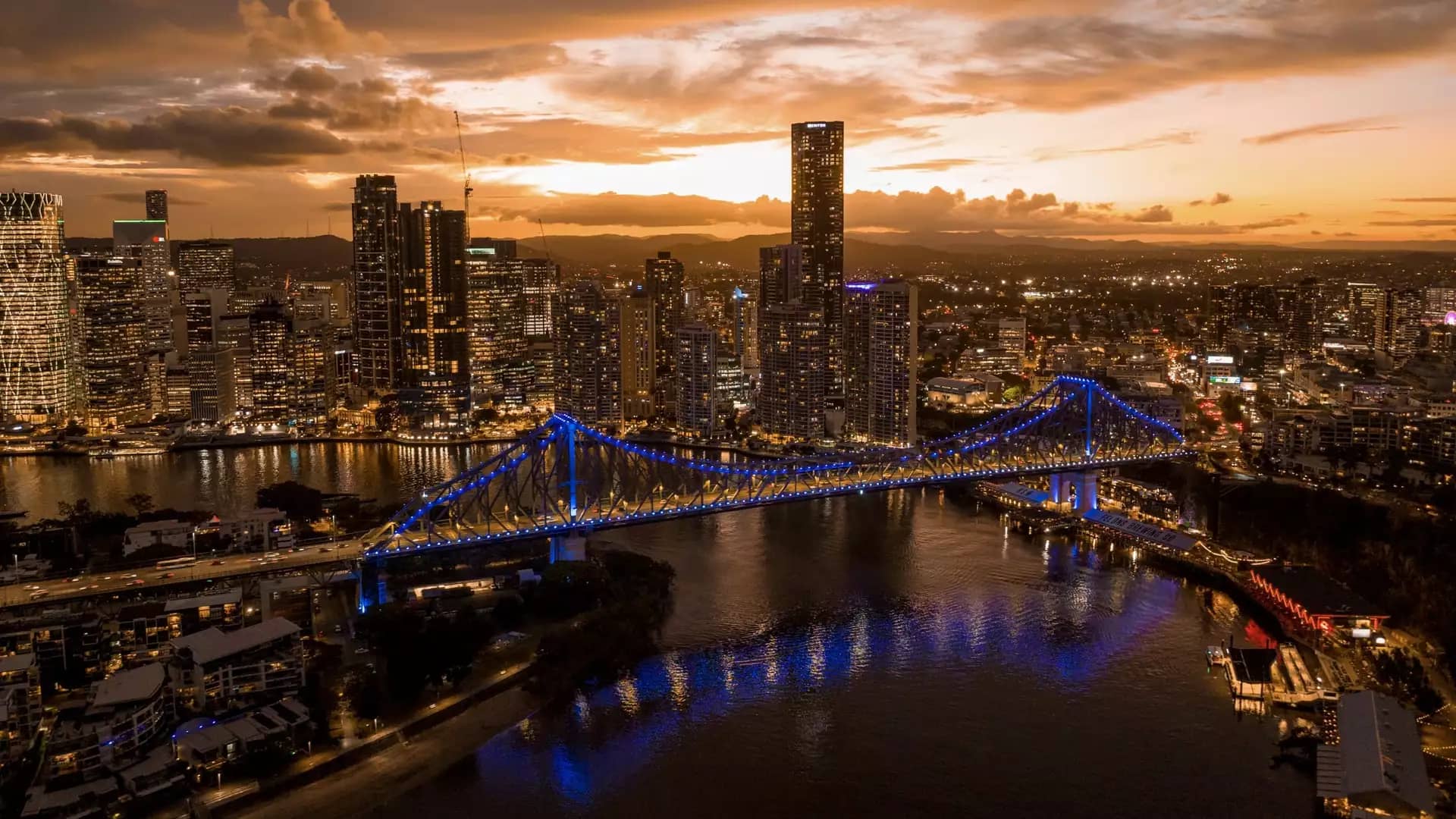 A dramatic wide aerial of the Brisbane CBD at sunset, featuring the illuminated Story Bridge spanning the river as the city lights begin to glow.