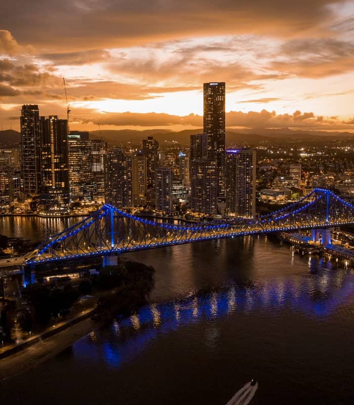 A dramatic wide aerial of the Brisbane CBD at sunset, featuring the illuminated Story Bridge spanning the river as the city lights begin to glow.