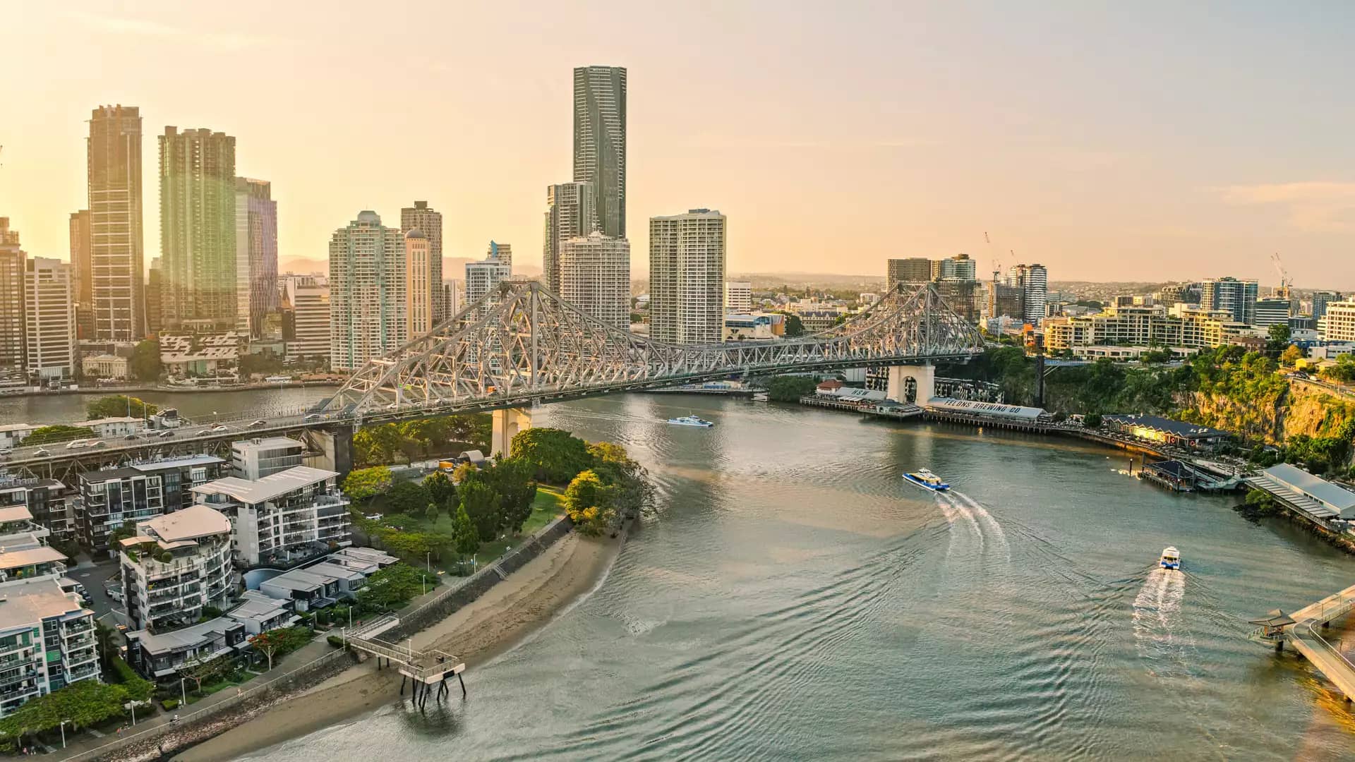 An aerial view of a wide river with several boats leaving white wakes, passing under a large steel bridge toward a city of tall buildings under a hazy yellow sky.