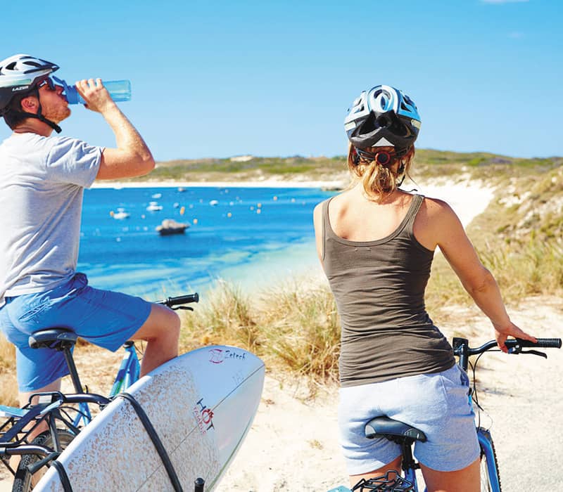 A man and woman in helmets sitting on bicycles overlooking a bright blue bay and beach at Rottnest Island.