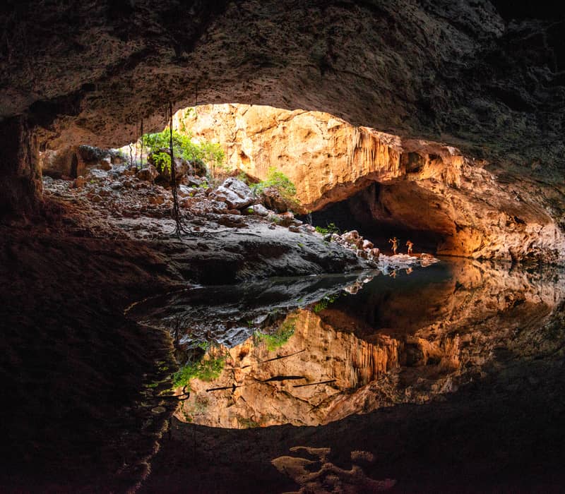Two adventurers wade through the permanent freshwater pools of Tunnel Creek, with the ancient limestone formations of the Napier Range reflecting in the still water.