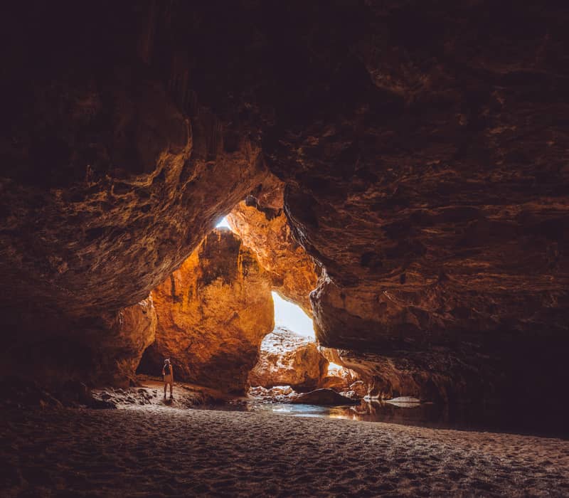 A traveler stands inside the vast limestone cavern of the Dimalurru (Tunnel Creek) cave system, where sunlight filters through a collapsed section of the roof onto the sandy floor.