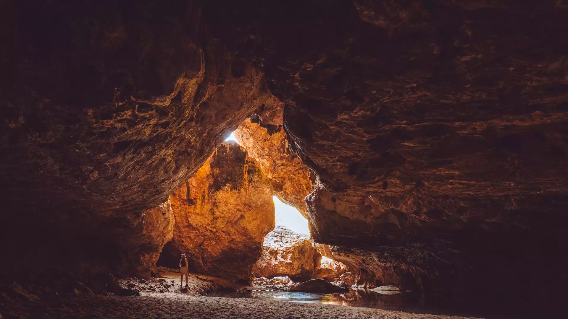 A traveler stands inside the vast limestone cavern of the Dimalurru (Tunnel Creek) cave system, where sunlight filters through a collapsed section of the roof onto the sandy floor.