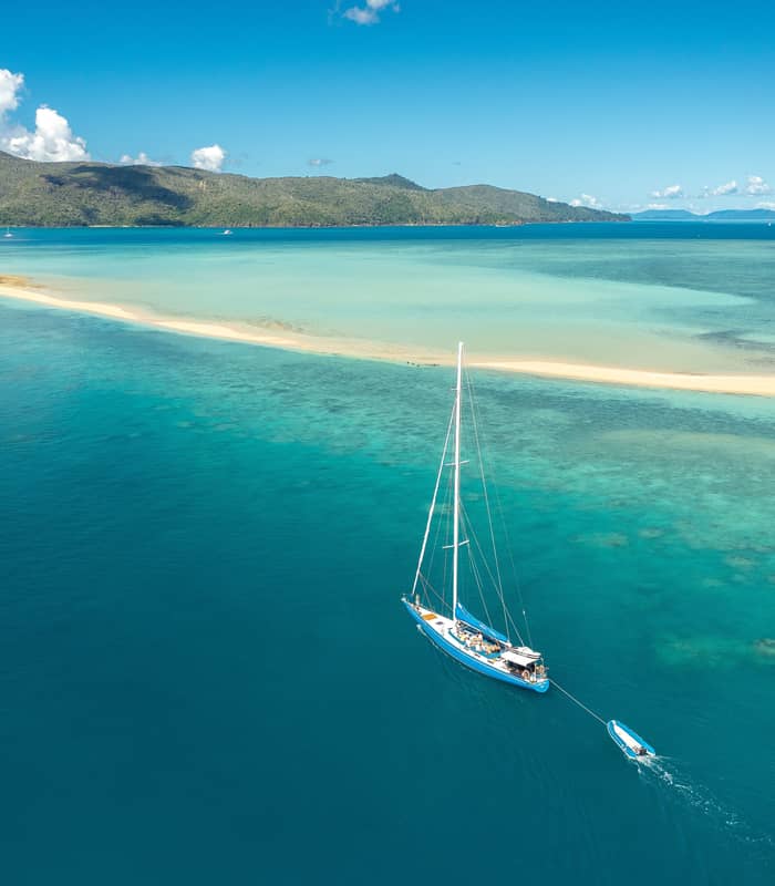A high-angle aerial view of a large blue sailboat anchored in crystal-clear water next to the long, narrow white sandspit of Langford Island in the Whitsundays.