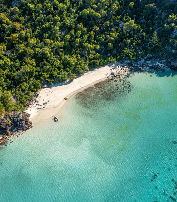 Looking straight down at a pristine white sand beach on a Whitsunday Island, where a small boat is pulled up to the shore near a dense, green tropical forest.